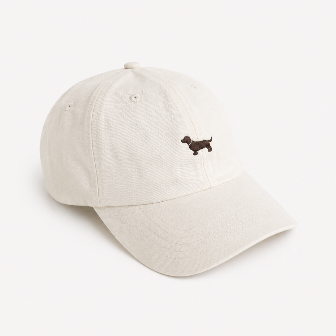 An off-white cotton baseball cap featuring a small embroidered brown dachshund on the front, set against a clean, solid off-white background.