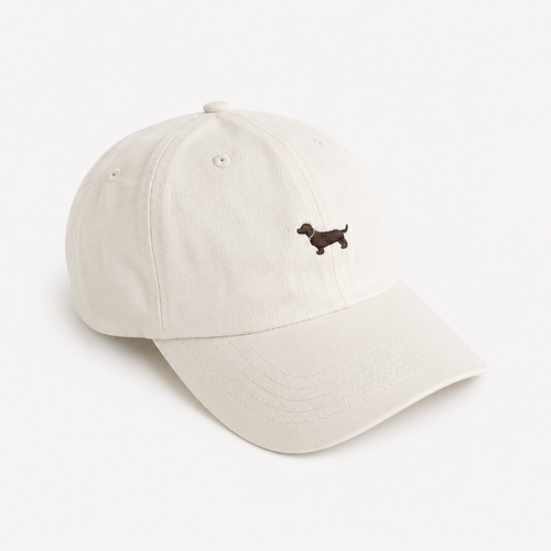 An off-white cotton baseball cap featuring a small embroidered brown dachshund on the front, set against a clean, solid off-white background.