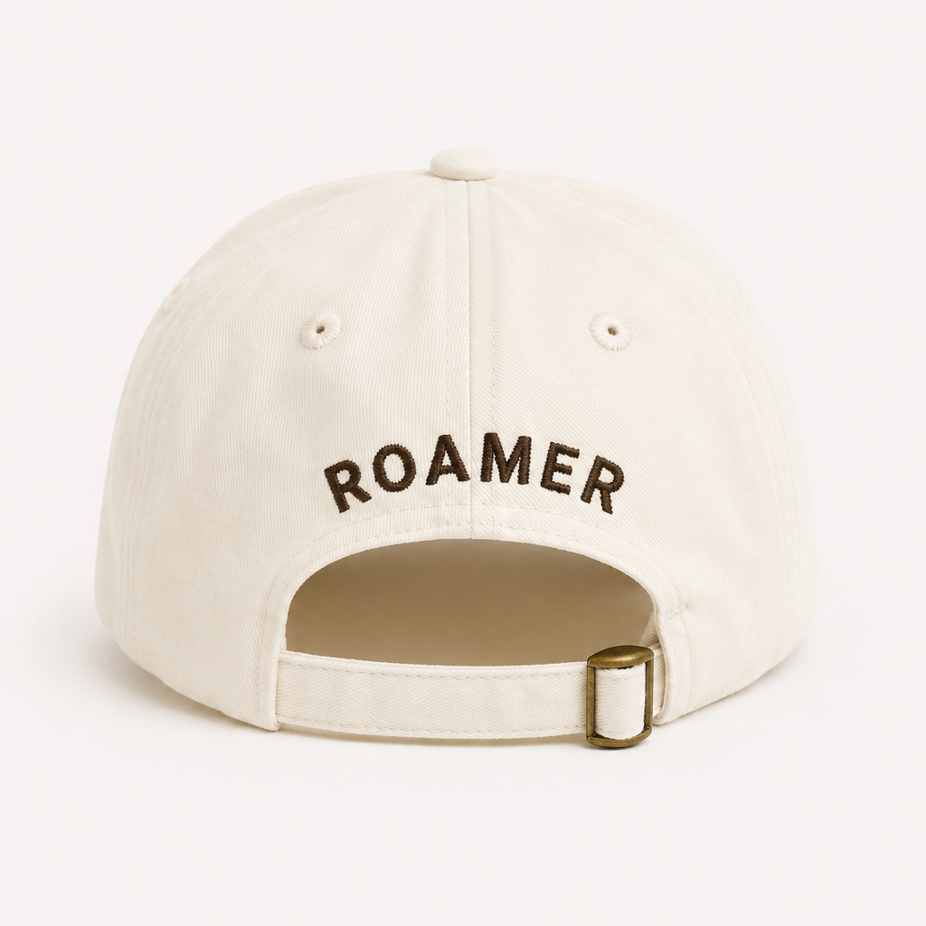 An off-white baseball cap shown from the back, featuring the word 'ROAMER' embroidered in black arched text and an adjustable strap with a gold-tone metal buckle.